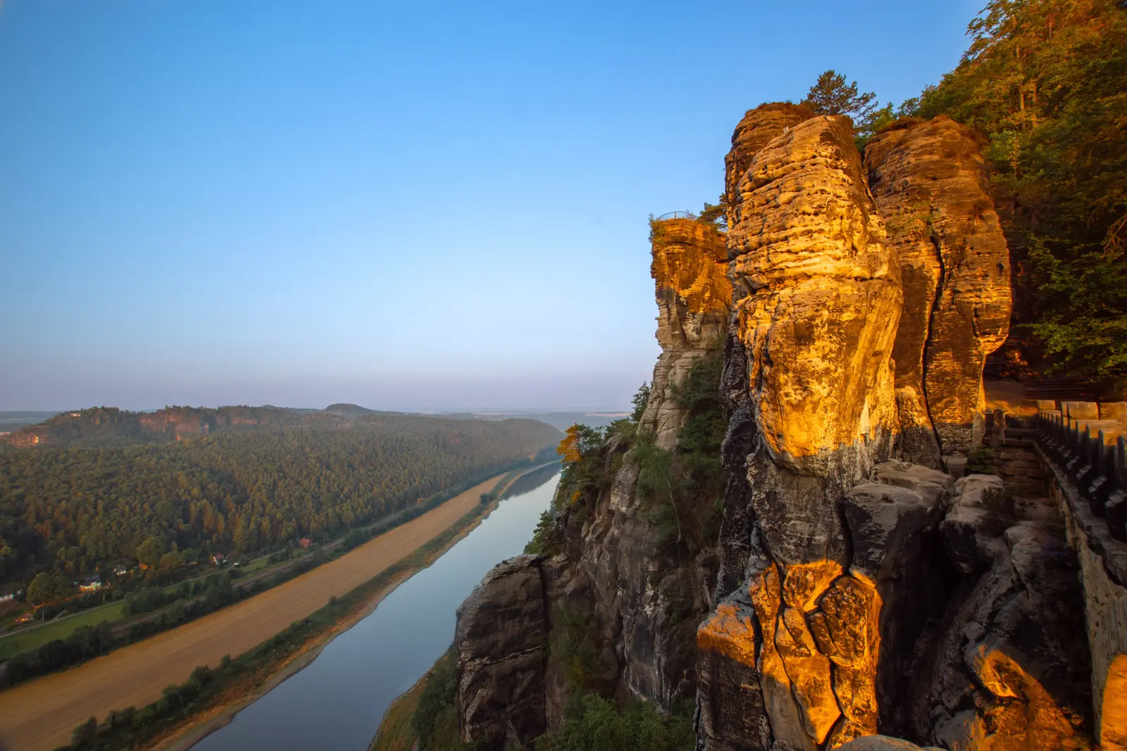 Blick aus der Höhe auf die Elbe, die sich durch das bewaldete Tal windet; rechts eine sonnenbeschienene Felsnadel.