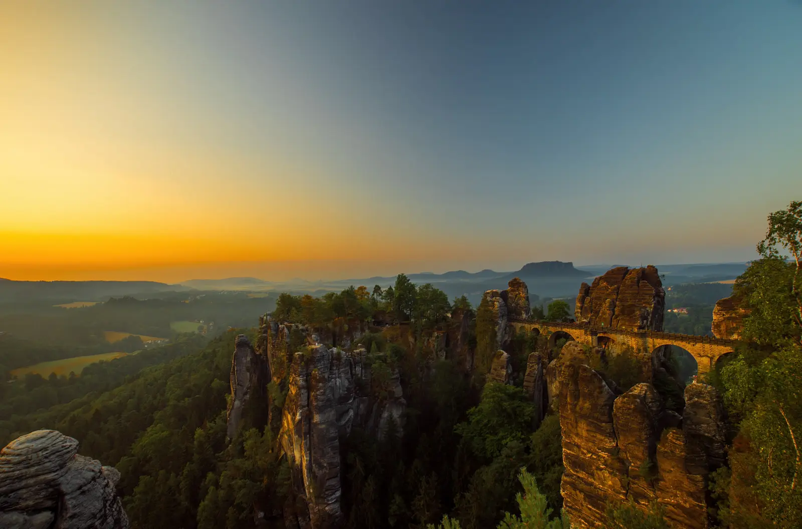 Weiter Blick über das Elbtal kurz nach Sonnenaufgang, die Basteibrücke von der Seite, am Horizont ein Tafelberg.