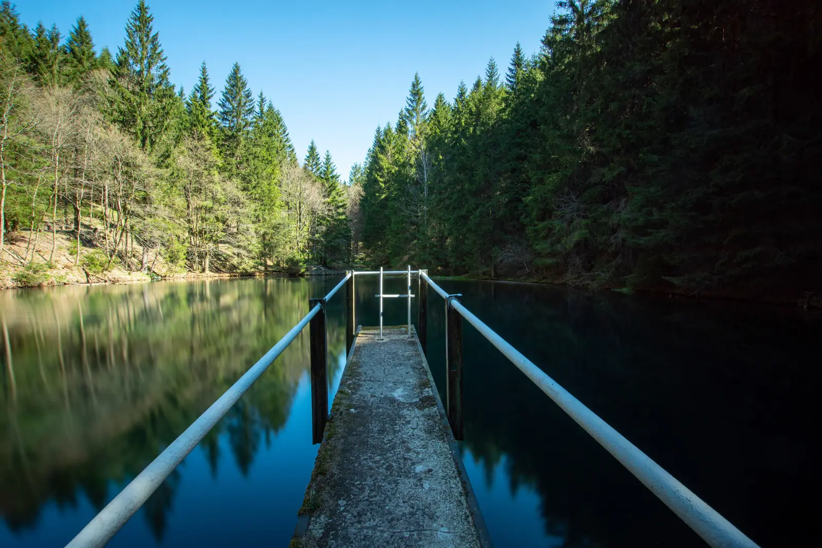 Schmaler Steg auf eine Talsperre hinaus, dunkles Wasser zwischen bewaldeten Hängen.