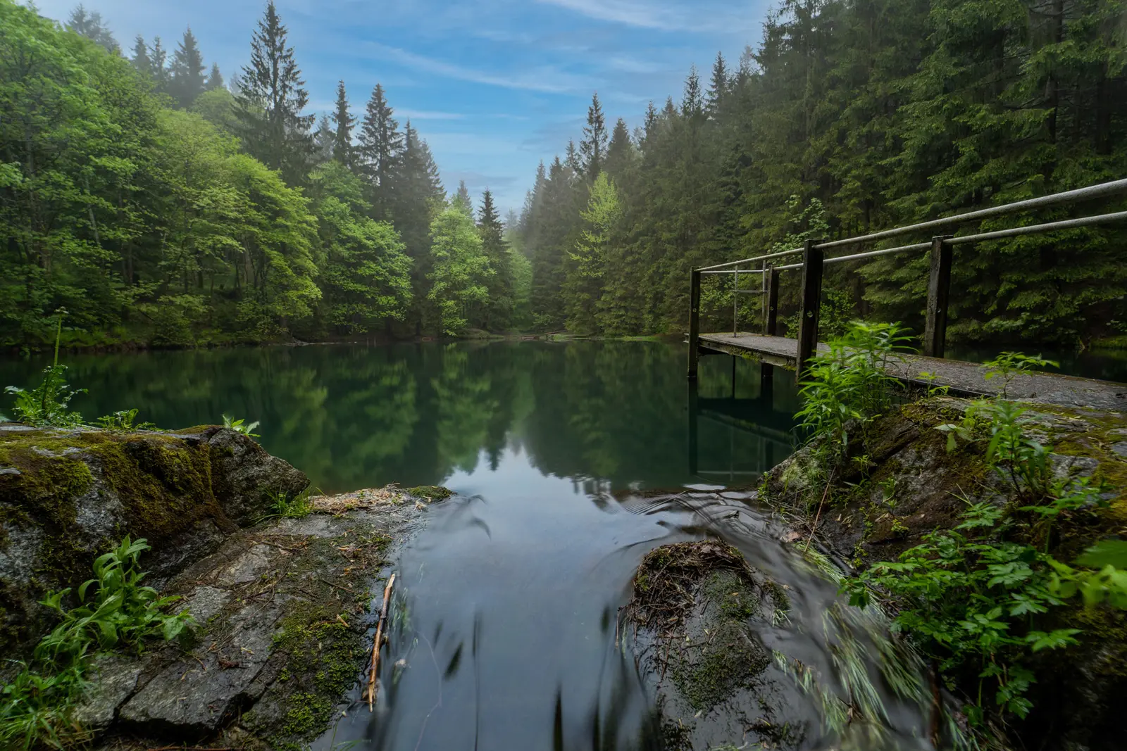 Kleiner Wasserfall an einem Waldsee im Sommer, rechts das Geländer eines Stegs.